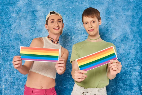 Lächelnde queere Personen in lässiger Kleidung mit lgbt-Fahnen und Blick in die Kamera während der stolzen Monatsfeier auf fleckigem und strukturiertem blauem Hintergrund — Stockfoto