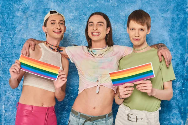 Positivo y tatuado hombre gay abrazando amigos sosteniendo banderas lgbt y mirando a la cámara durante la celebración del mes de orgullo sobre fondo azul moteado - foto de stock