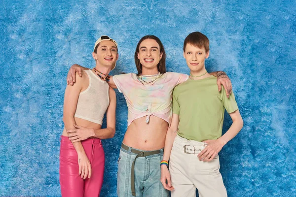 Улыбающийся длинноволосый педик, обнимающий друзей-гомосексуалистов и смотрящий в камеру во время празднования месяца гей-гордости на пестром синем фоне — стоковое фото