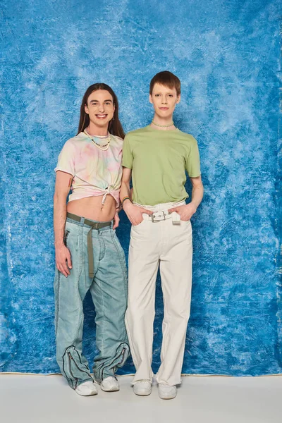 Полная длина улыбающихся друзей-геев в непринужденной одежде, позирующих и смотрящих в камеру во время празднования месяца гордости на пестром синем фоне — стоковое фото