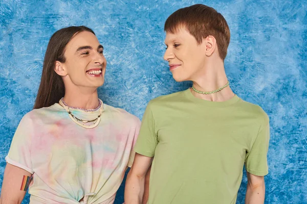 Portrait of cheerful queer friends in casual clothes looking at each other during lgbt pride month celebration on mottled textured blue background — Stock Photo