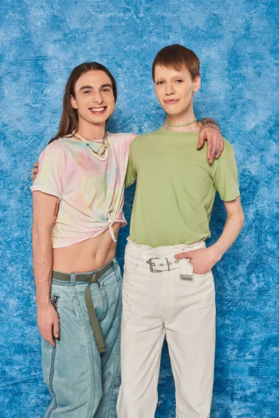 Молодые друзья-гомосексуалисты позируют и обнимаются, глядя в камеру во время празднования месяца гордости на пестром синем фоне с пятнами — стоковое фото