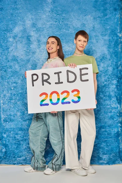 Toute la longueur des amis queer joyeux regardant la caméra tout en tenant la plaque avec fierté 2023 lettrage pendant la célébration du mois lgbt sur fond bleu marbré — Photo de stock