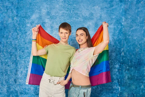 Alegre y joven homosexual amigos sosteniendo bandera lgbt juntos y mirando a la cámara durante el mes de orgullo celebración de la comunidad sobre fondo azul moteado - foto de stock