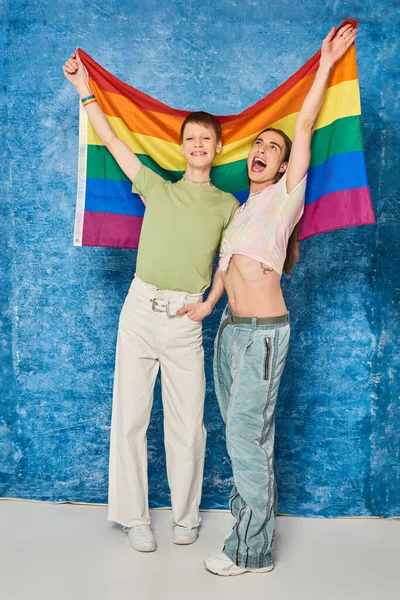 Longitud completa de la comunidad homosexual emocionada con ropa casual sosteniendo la bandera lgbt mientras celebran el mes de orgullo y de pie juntos sobre fondo azul moteado - foto de stock