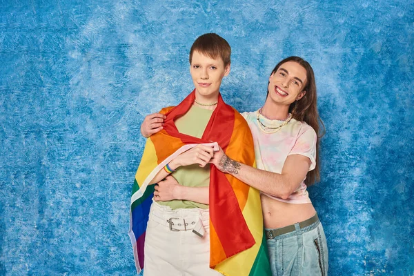 Smiling tattooed gay man in casual clothes hugging young friend with lgbt flag and looking at camera during pride month celebration on mottled blue background — Stock Photo