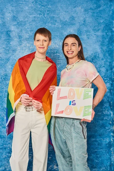 Unbekümmerte queere Gemeinschaft mit lgbt-Fahne, die ein Plakat mit Liebe hält, ist Liebe Schriftzug und blickt in die Kamera, während sie Stolz Monat auf fleckigem blauem Hintergrund feiert — Stockfoto