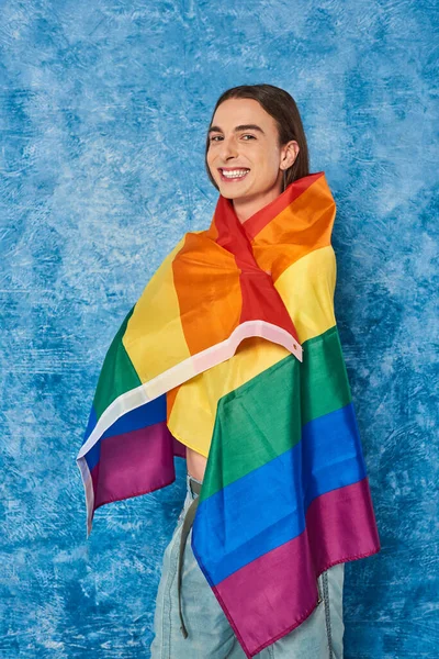 Positivo homem homossexual de cabelos longos olhando e câmera e envolvendo em lgbt bandeira durante orgulho comunidade mês celebração no fundo azul manchado — Fotografia de Stock