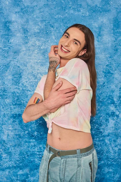 Carefree long haired and tattooed queer person smiling at camera and posing during lgbt month celebration on mottled and textured blue background — Stock Photo