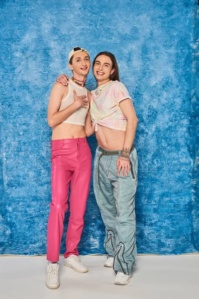 Full length of positive gay friends in stylish outfits hugging and looking at camera during lgbt pride month celebration on textured blue background — Stock Photo