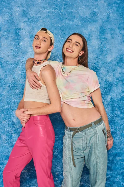 Portrait of smiling and stylish queer friends hugging and looking at camera together during lgbt pride month celebration on textured blue background — Stock Photo