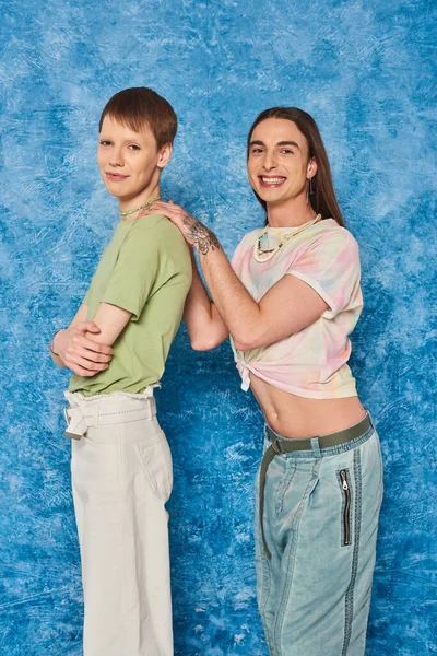 Sonriente hombre gay tatuado y de pelo largo abrazando a un amigo queer cruzando brazos durante la celebración de la polilla del orgullo lgbt sobre un fondo azul texturizado - foto de stock