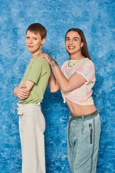 Souriant tatoué et aux cheveux longs gay homme étreignant queer ami croisement bras pendant lgbt fierté papillon de nuit célébration sur texturé fond bleu — Photo de stock