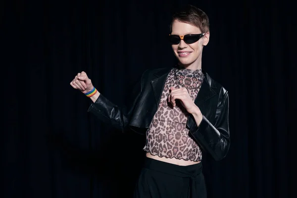 Positiver und stilvoller homosexueller Mann in Sonnenbrille, Lederjacke und Bluse mit Animal Print posiert während der lgbt Pride Monatsparty isoliert auf schwarz — Stockfoto