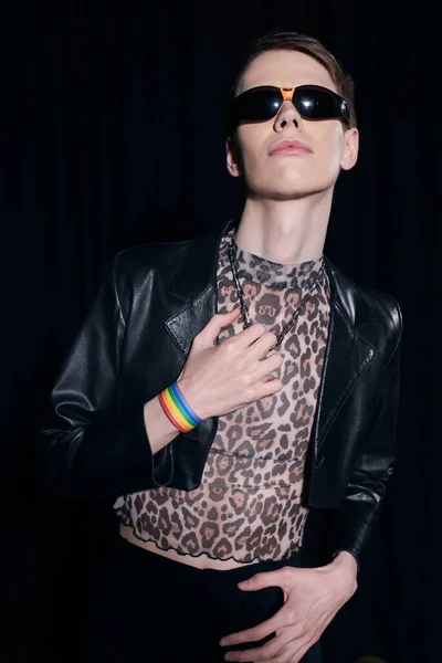 Retrato de persona queer de moda en gafas de sol, blusa con estampado animal y chaqueta de cuero posando durante la celebración del mes lgbt aislado en negro - foto de stock