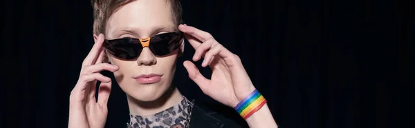 Portrait of trendy nonbinary person with lgbt flag on bracelet touching sunglasses during pride community month celebration isolated on black, banner — Stock Photo