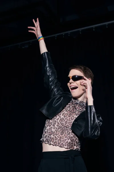 Carefree nonbinary person in stylish sunglasses, leather jacket and blouse with animal print celebrating pride month on black background — Stock Photo