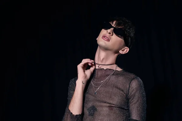 Alla moda gay uomo in occhiali da sole e frizzante top toccare collana durante lgbt comunità e orgoglio mese partito isolato su nero — Foto stock