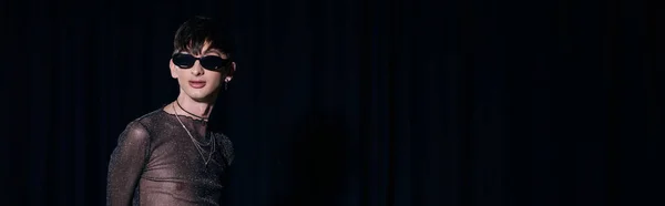 Persona alla moda non binaria in occhiali da sole e scintilla alto in piedi durante lgbt orgoglio mese celebrazione isolato su nero con spazio copia, banner — Foto stock