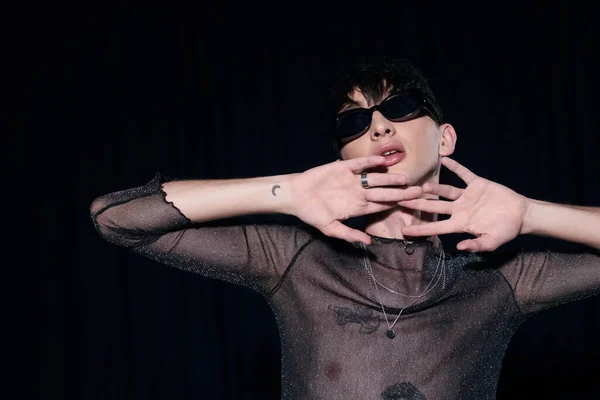 Tätowierter und modischer schwuler Mann mit Sonnenbrille und funkelndem Top, der das Gesicht während der lgbt-Monatsfeier berührt, isoliert auf schwarz — Stockfoto