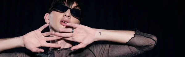 Портрет молодого, татуированного и модного гея в солнцезащитных очках и зажигательный топ, позирующий во время празднования месяца гордости, изолированного на черном, баннере — стоковое фото