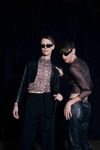 Модный гей в сверкающих топах и солнцезащитных очках, позирующий рядом с молодым другом во время празднования месяца гордости, изолированного на черном — стоковое фото
