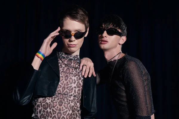 Portrait of fashionable queer persons in sunglasses and party outfits posing and looking at camera during lgbt pride month celebration isolated on black — Stock Photo