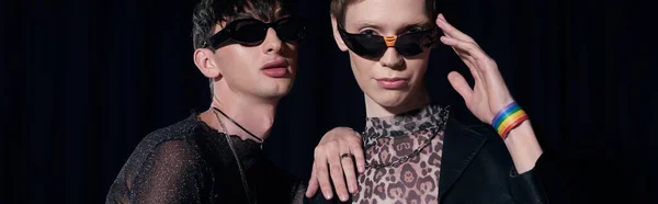 Стильный гей в солнцезащитных очках трогает плечо друга, глядя в камеру во время празднования долгого месяца, изолированного на черном баннере — стоковое фото
