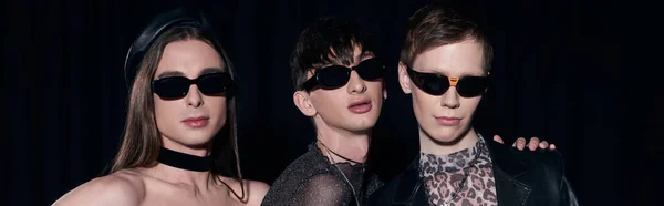 Portrait de couple homosexuel à la mode en vêtements de fête et lunettes de soleil posant pendant la célébration du mois de fierté lgbt isolé sur noir, bannière — Photo de stock