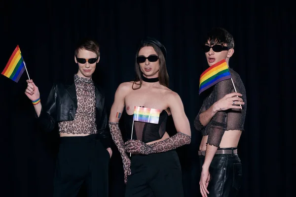 Молодые и стильные друзья-гомосексуалисты в солнцезащитных очках и костюмах для вечеринок с флагами lgbt во время празднования месяца гордости, изолированного на черном — стоковое фото