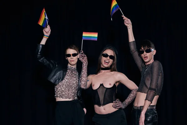 Carefree and fashionable homosexual friends in sunglasses and party clothes holding rainbow flags during pride month celebration isolated on black — Stock Photo