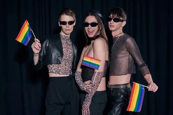 Fashionable and positive homosexual friends in party outfits and sunglasses posing with lgbtq flags during pride month celebration on black background — Stock Photo