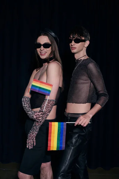 Fashionable homosexual friends in party outfits and sunglasses holding rainbow flags and posing during lgbt flag month celebration isolated on black — Stock Photo