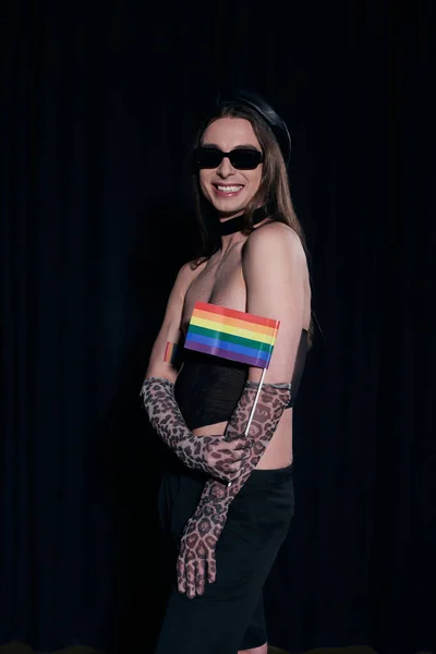 Unbekümmerter und langhaariger schwuler Mann mit Sonnenbrille, sexy Oberteil und Handschuhen, der die lgbt-Fahne während der Pride Community Monatsparty hält, isoliert auf schwarz — Stockfoto