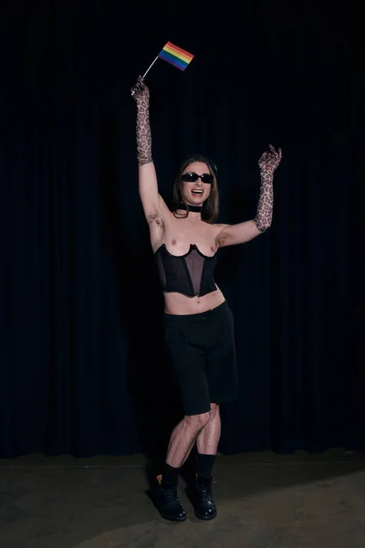 Lunghezza completa della persona queer eccitata e alla moda in occhiali da sole e corsetto sexy con bandiera lgbt durante la celebrazione del mese dell'orgoglio su sfondo nero — Foto stock