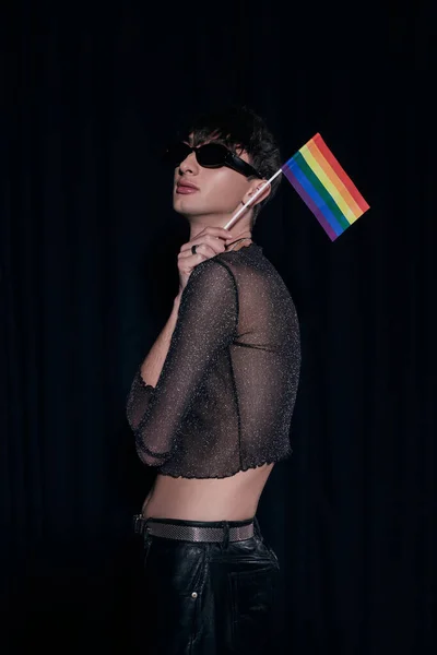 Hombre homosexual de moda y joven en gafas de sol y la parte superior brillante celebración de la bandera del arco iris durante lgbt orgullo mes aislado en negro - foto de stock