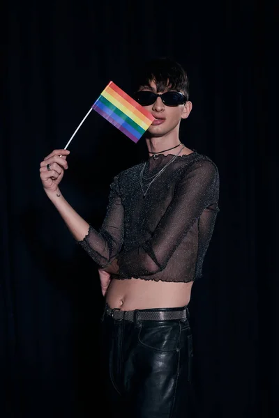 Trendy homosexual young man in sunglasses and shiny top biting lgbt flag and posing during pride community month celebration isolated on black — Stock Photo