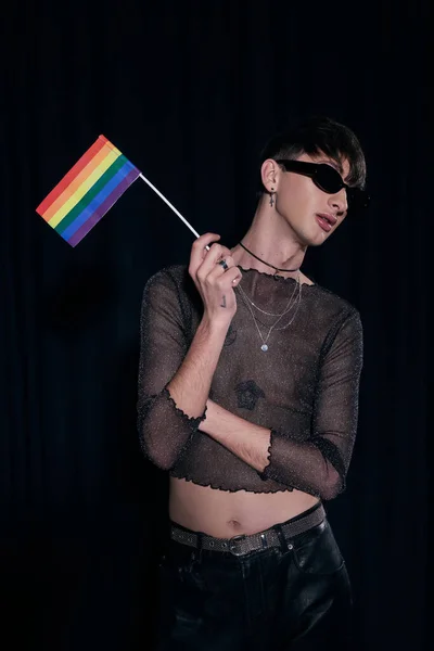 Junge und modische queere Person mit Sonnenbrille und glänzendem Oberteil, die die lgbt-Fahne hält, während sie während der stolzen Monatsfeier der Gemeinschaft isoliert auf schwarz posiert — Stockfoto