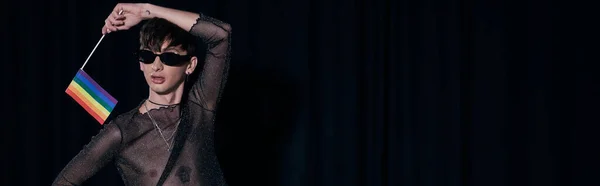 Junge und stylische nichtbinäre Person in Sonnenbrille und glänzendem Oberteil mit Regenbogenfahne während der Stolz-Gemeinschaftsfeier isoliert auf schwarz, Banner — Stockfoto