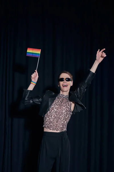 Excited nonbinary person in stylish outfit and sunglasses holding lgbt flag during party and celebration of pride month on black background — Stock Photo