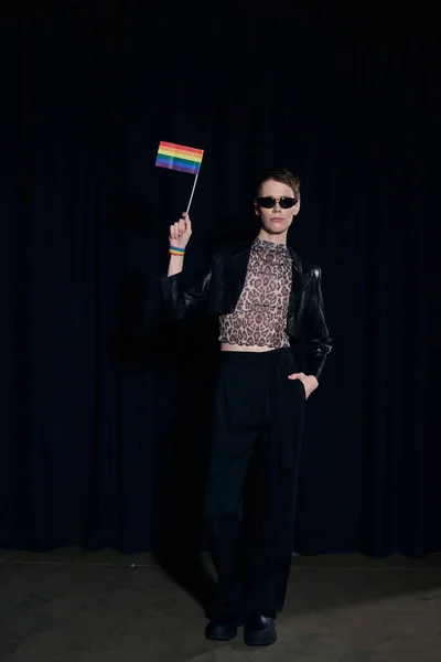 Полная длина стильный небинарный человек в костюме партии и солнцезащитные очки, держа радужный флаг и позируя во время lgbt гордости месяц празднования на черном фоне — стоковое фото