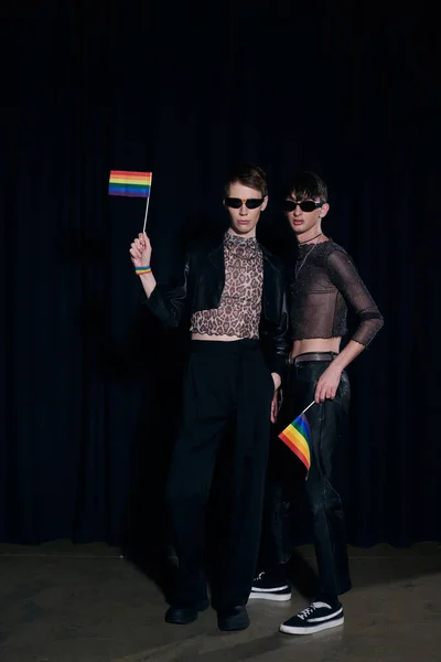 Full length of fashionable gay friends in sunglasses and party outfits holding rainbow lgbt flags during pride community month celebration on black background — Stock Photo