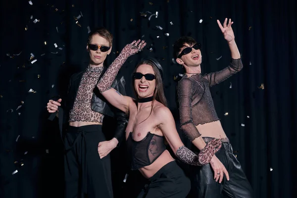 Trendy and positive nonbinary friends in sunglasses and party clothes posing under falling confetti during lgbt pride month celebration on black background — Stock Photo