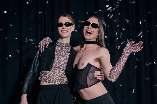 Sexy and cheerful homosexual friends in party outfits hugging while standing under falling confetti during lgbt pride month celebration on black background — Stock Photo