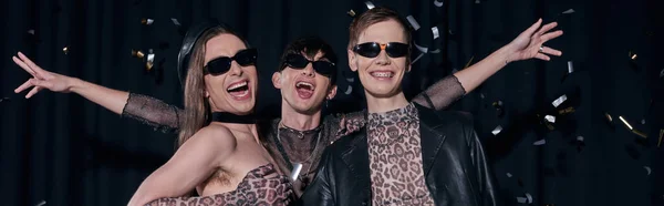 Група веселих і модних гомосексуальних друзів у партійному вбранні та сонцезахисних окулярах, що стоять під падінням конфеті під час святкування місяця lgbt на чорному тлі, банер — стокове фото