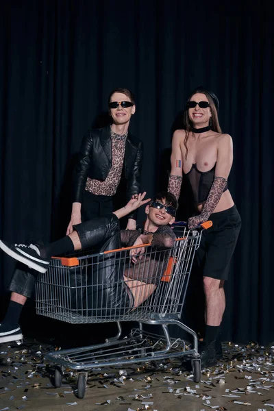 Smiling group of homosexual friends in stylish party outfits having fun with shopping cart during lgbt month celebration on black background — Stock Photo