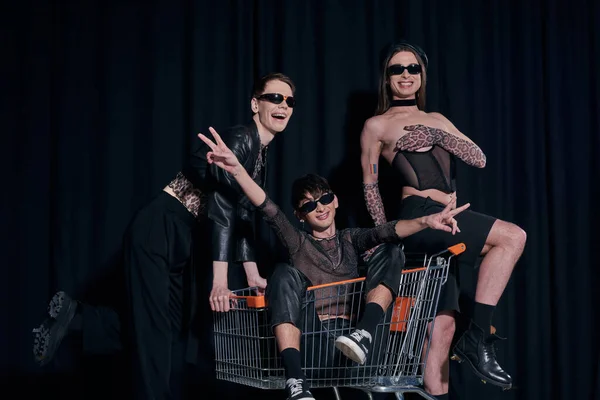 Smiling and stylish group of homosexual friends in sunglasses having fun with friend in shopping cart during pride month celebration on black background — Stock Photo