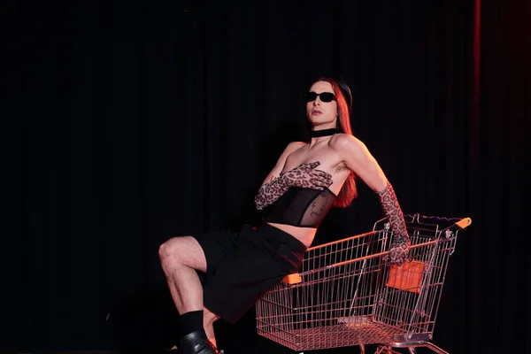 Fashionable homosexual man in sunglasses and sexy corset sitting on shopping cart during party and lgbt pride month celebration on black background — Stock Photo