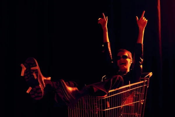 Aufgeregte nichtbinäre Person mit Sonnenbrille und stylischem Outfit, die mit den Fingern zeigt, während sie im Einkaufswagen sitzt, während lgbt Stolz Monatsfeier auf schwarzem Hintergrund mit rotem Licht — Stockfoto