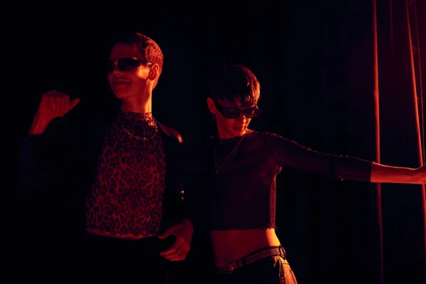 Amigos não binários na moda em roupas de festa e óculos de sol dançando juntos enquanto celebram o mês do orgulho lgbt no fundo preto com iluminação vermelha — Fotografia de Stock